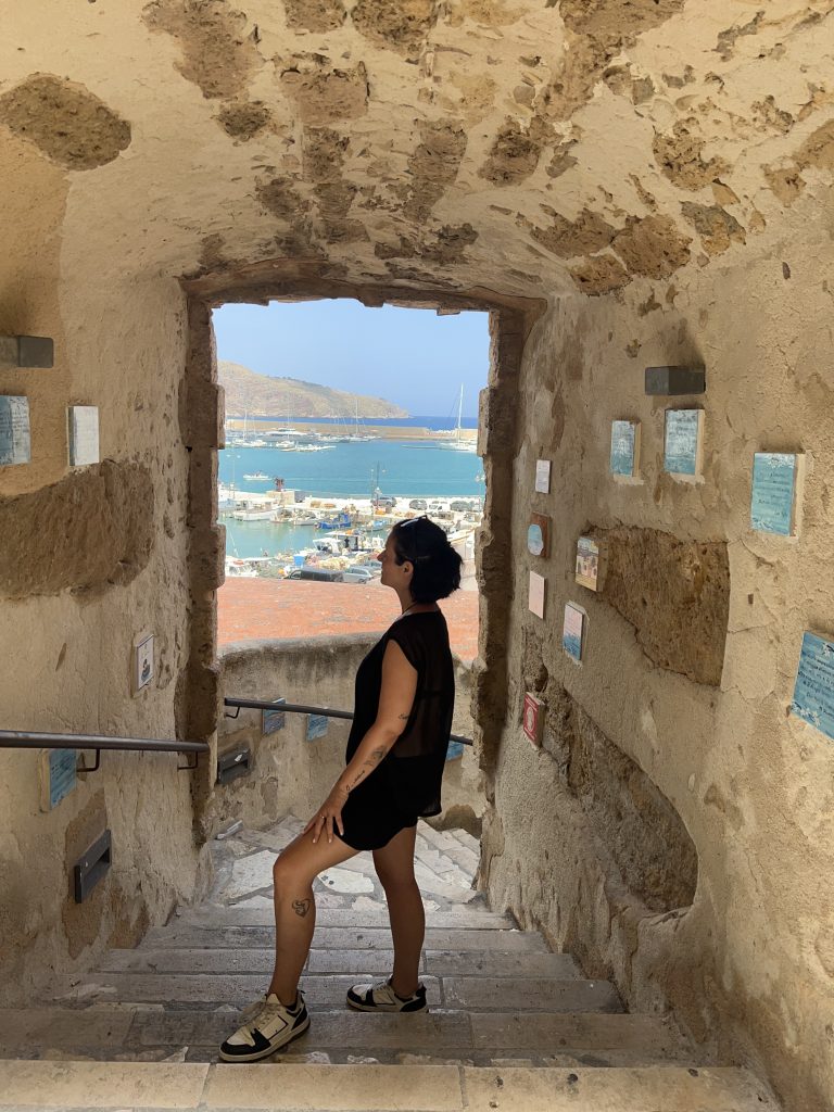 Simona osserva il porto di Castellammare del Golfo attraverso un passaggio storico in pietra
